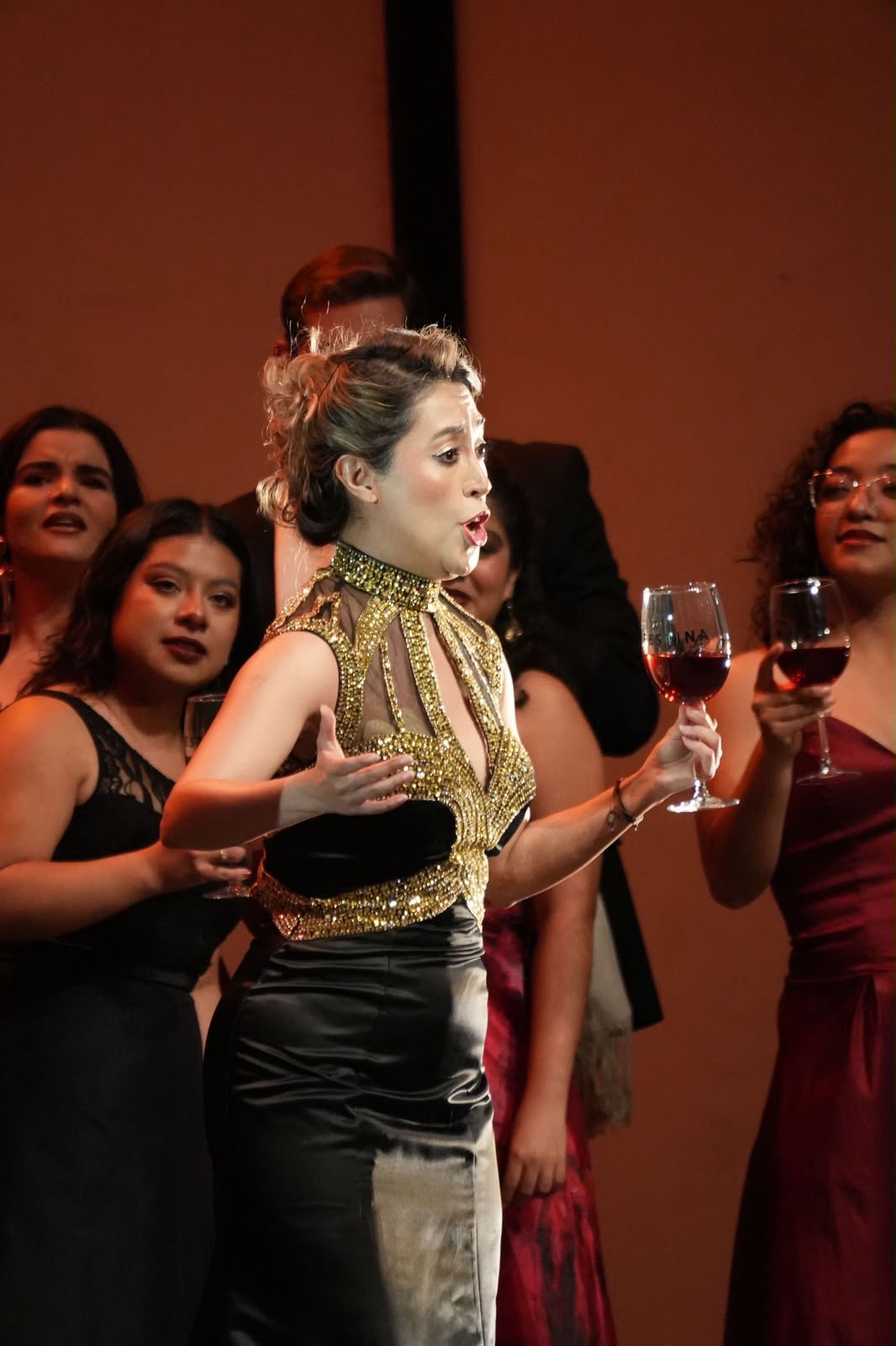 Escena de brindis operístico con vestuario dorado y copas en alto